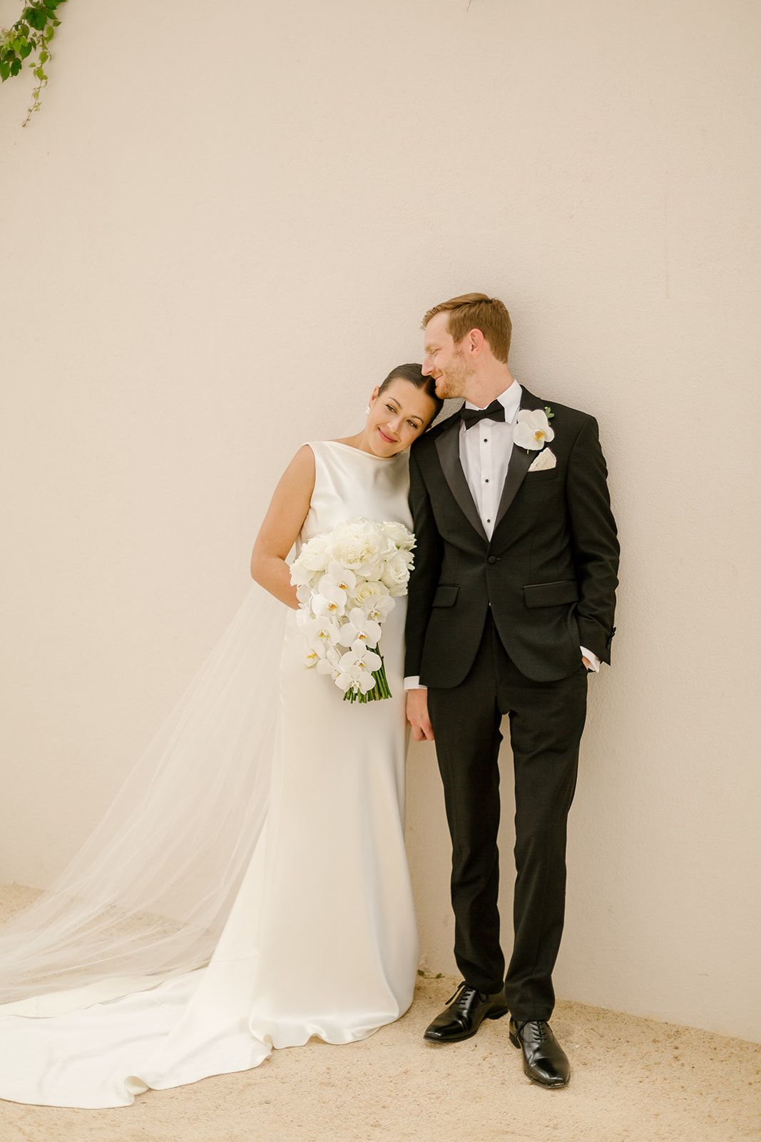 A couple in wedding attire embraces against a cream wall, with the bride holding an elegant white orchid bouquet.