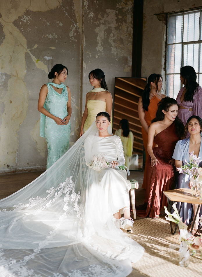 A wedding party gets ready in a rustic interior with bridesmaids in mint green and burgundy dresses tending to the bride's gown.