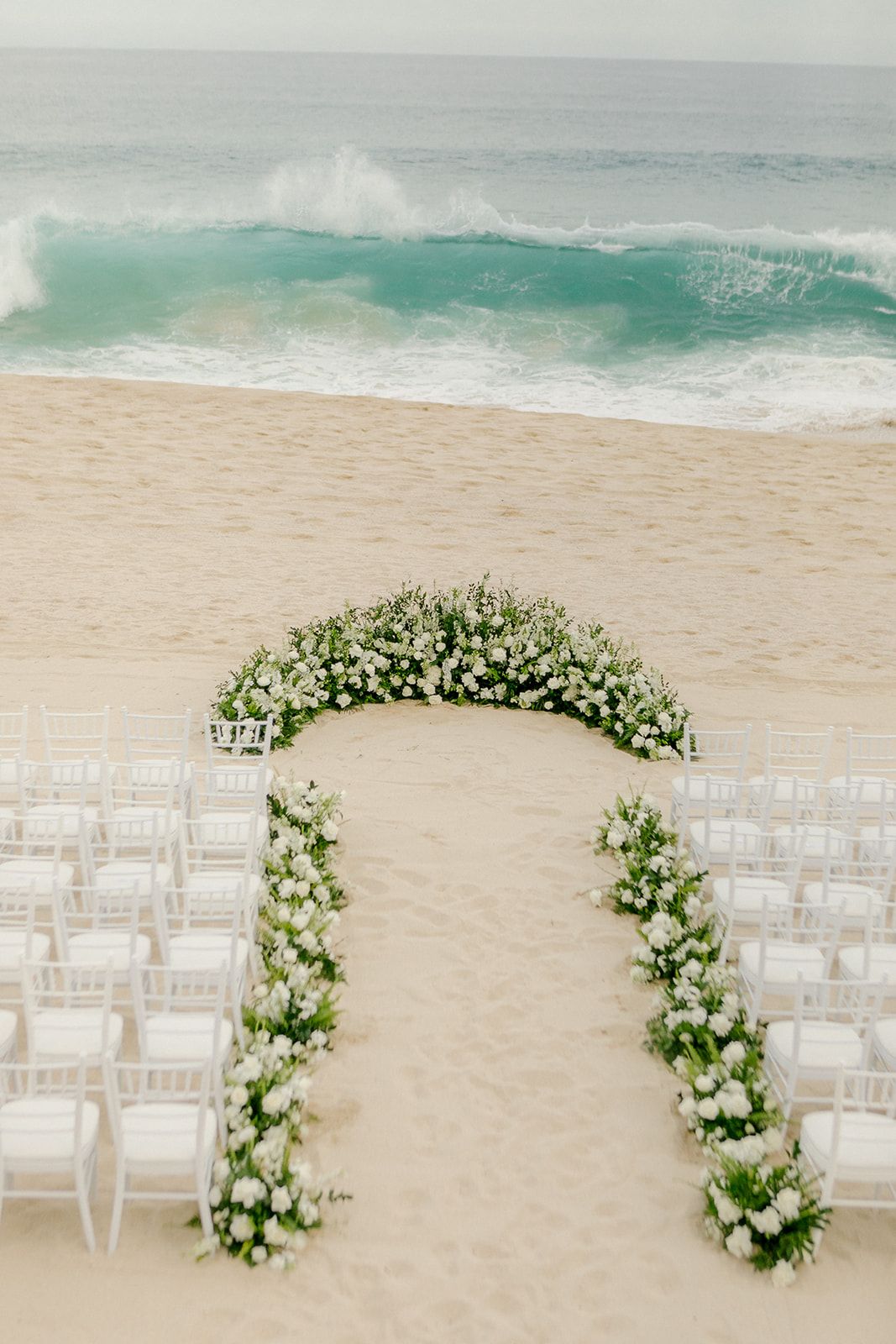 Wedding arch decorated with white flowers and chairs set up for a beach ceremony with turquoise waves crashing in background.