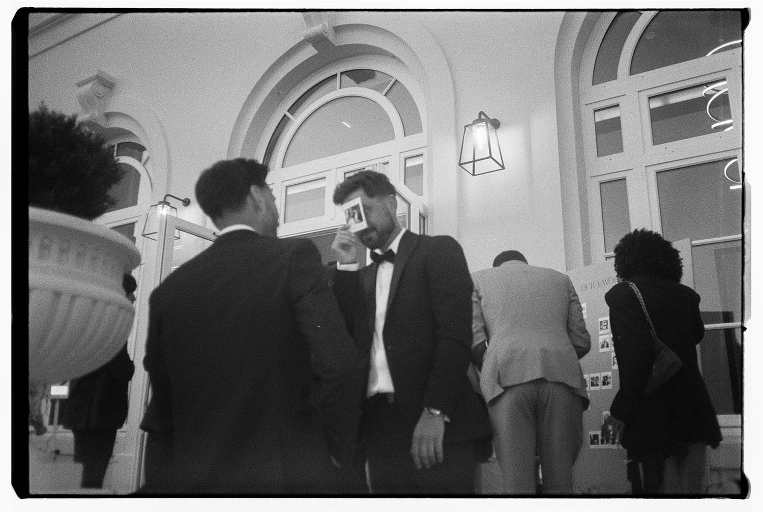 Black and white photograph showing people in formal attire gathered beneath an arched doorway with a hanging lamp.