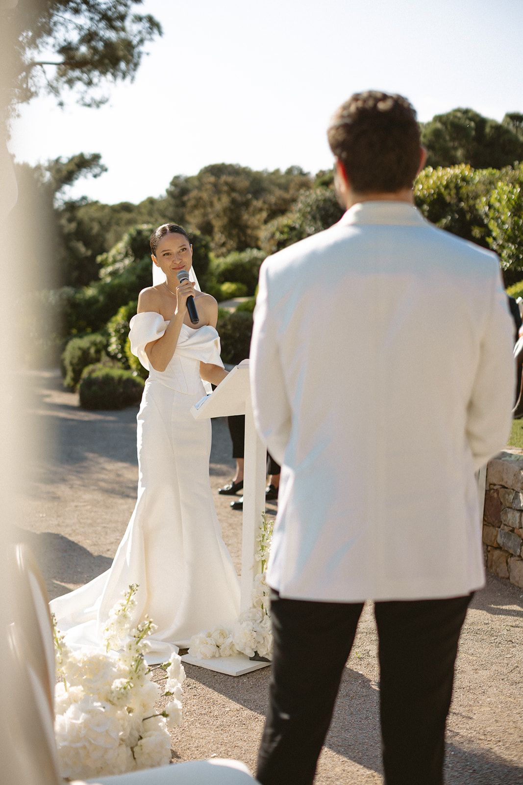 A couple in matching white formalwear share intimate moment during outdoor wedding ceremony in sunlit garden setting.