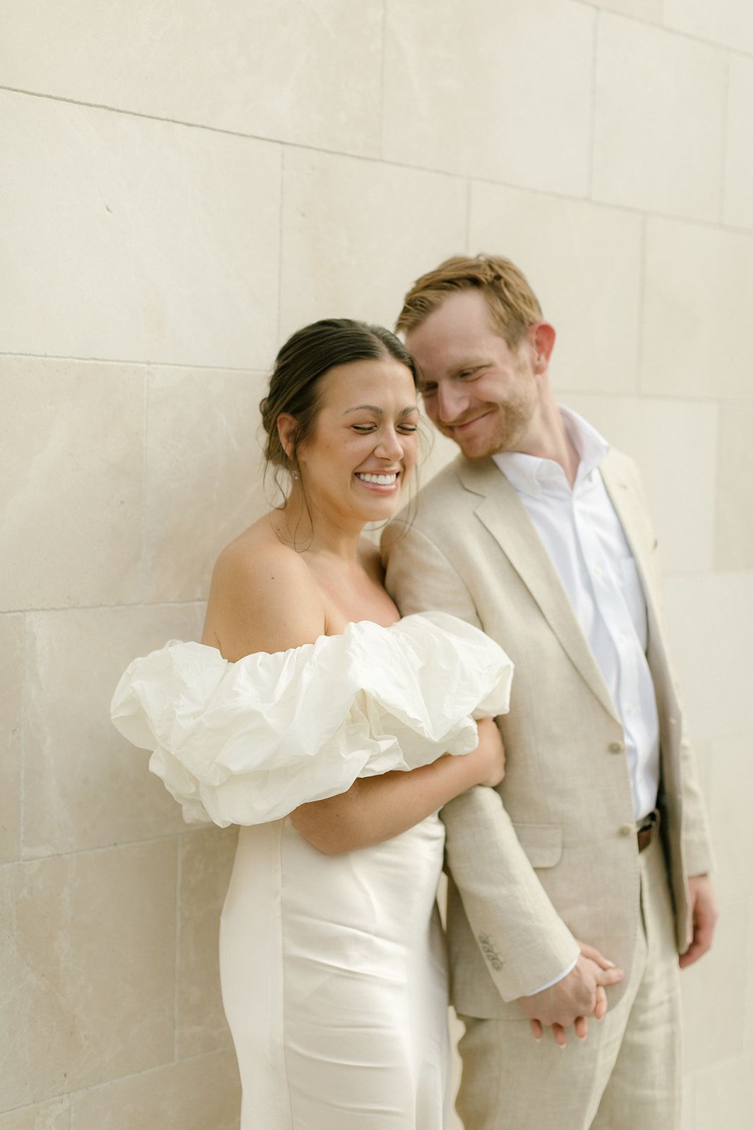 A joyful couple sharing an intimate moment against a light colored wall in wedding attire.