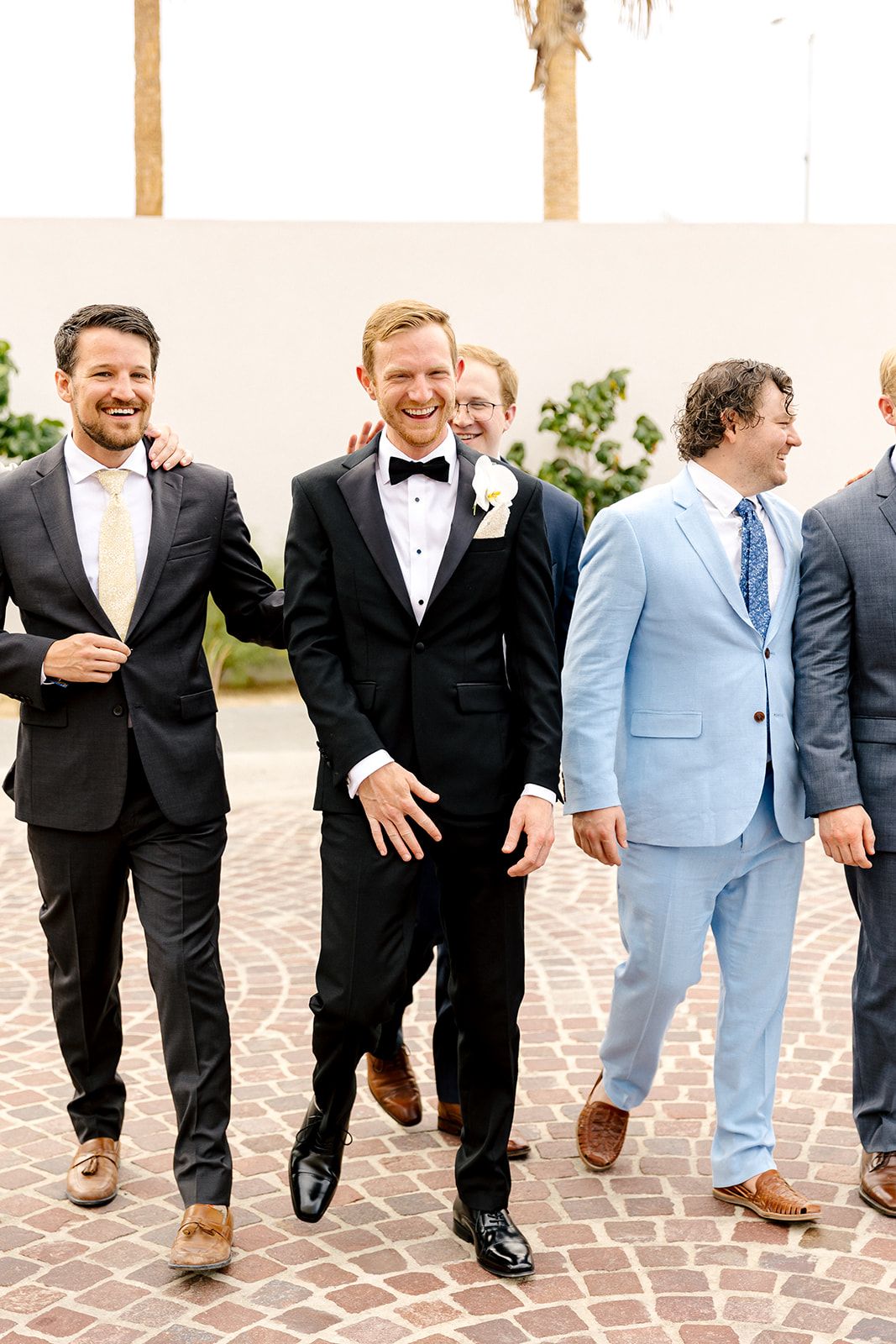 Wedding party including groomsmen in mixed formal attire walking together on brick pavement outside.