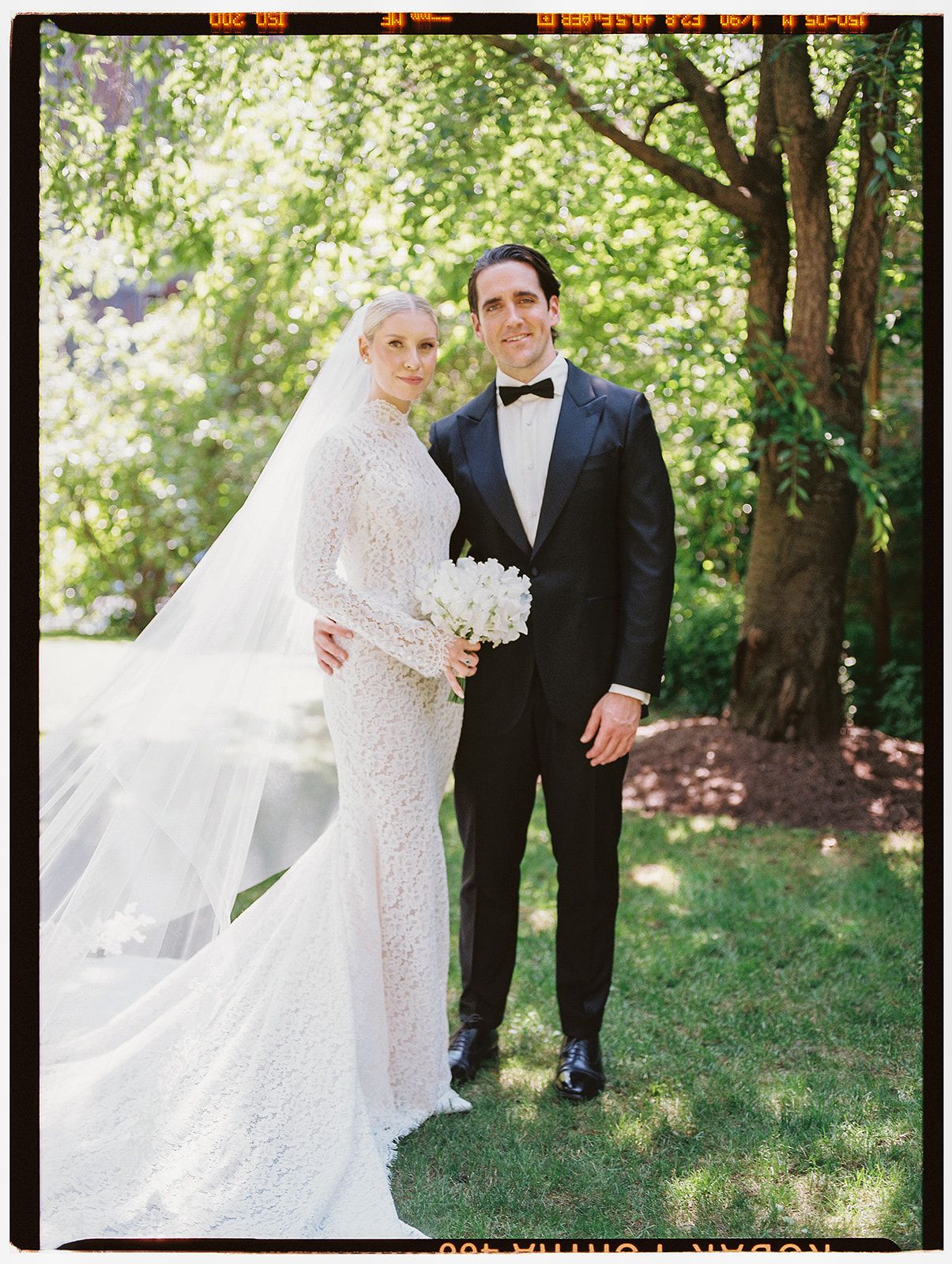 Wedding couple embraces outdoors among trees with bride's veil flowing in the breeze.