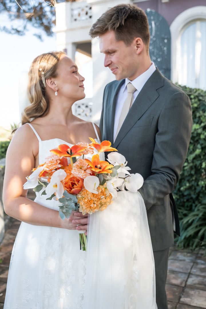 Newlyweds share tender moment with vibrant orange and white bouquet in outdoor garden setting.