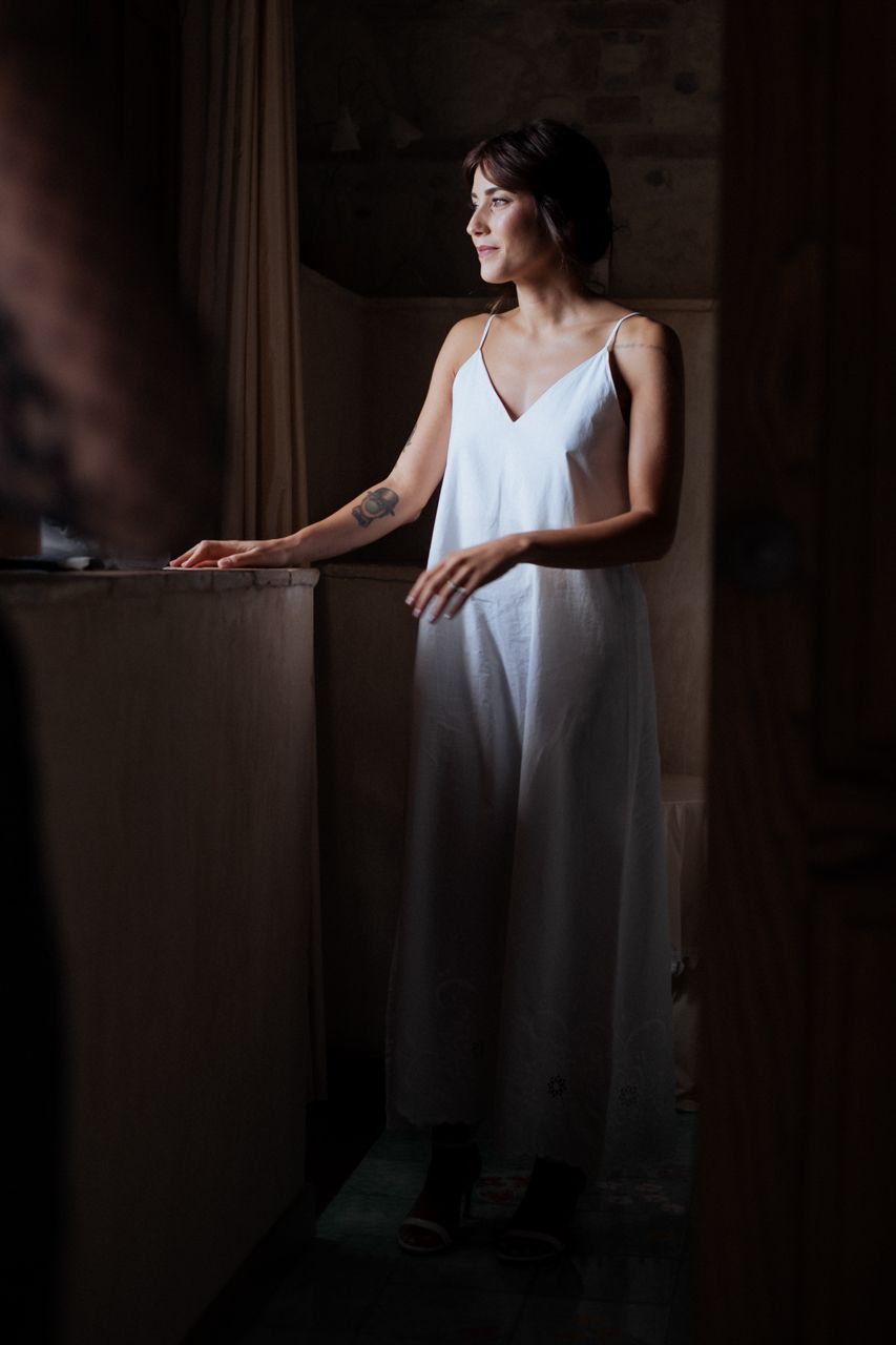 A figure in a white dress stands thoughtfully by a window in dramatic lighting.