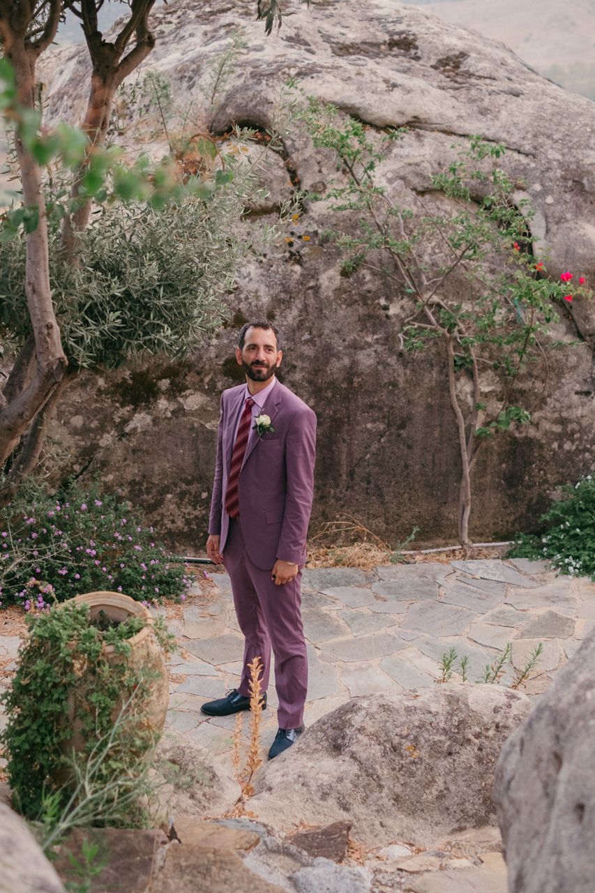 A person in a purple suit stands on rocky terrain surrounded by greenery and natural stone formations.