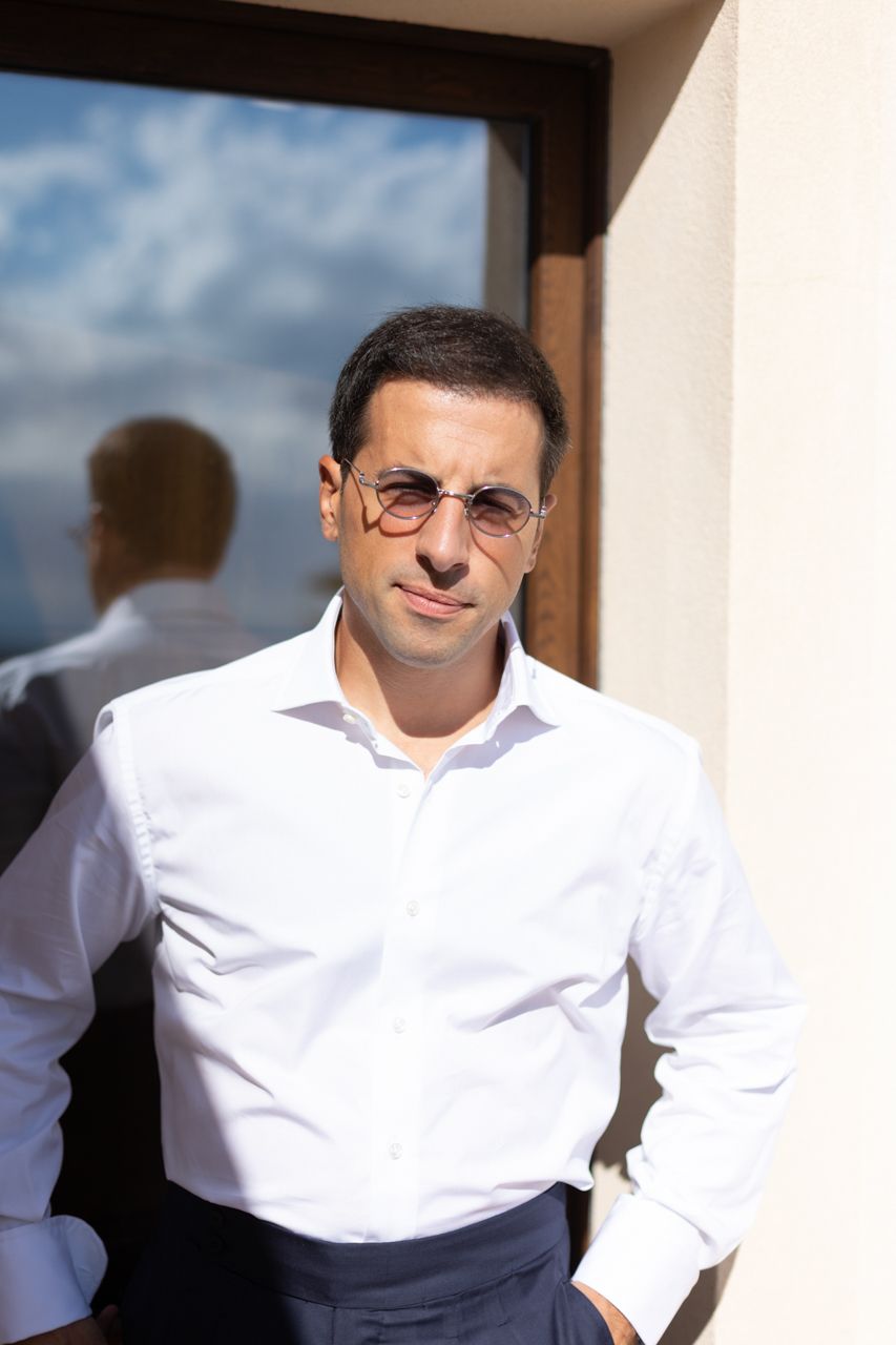 A person in a crisp white shirt and glasses poses against a sunlit wall.