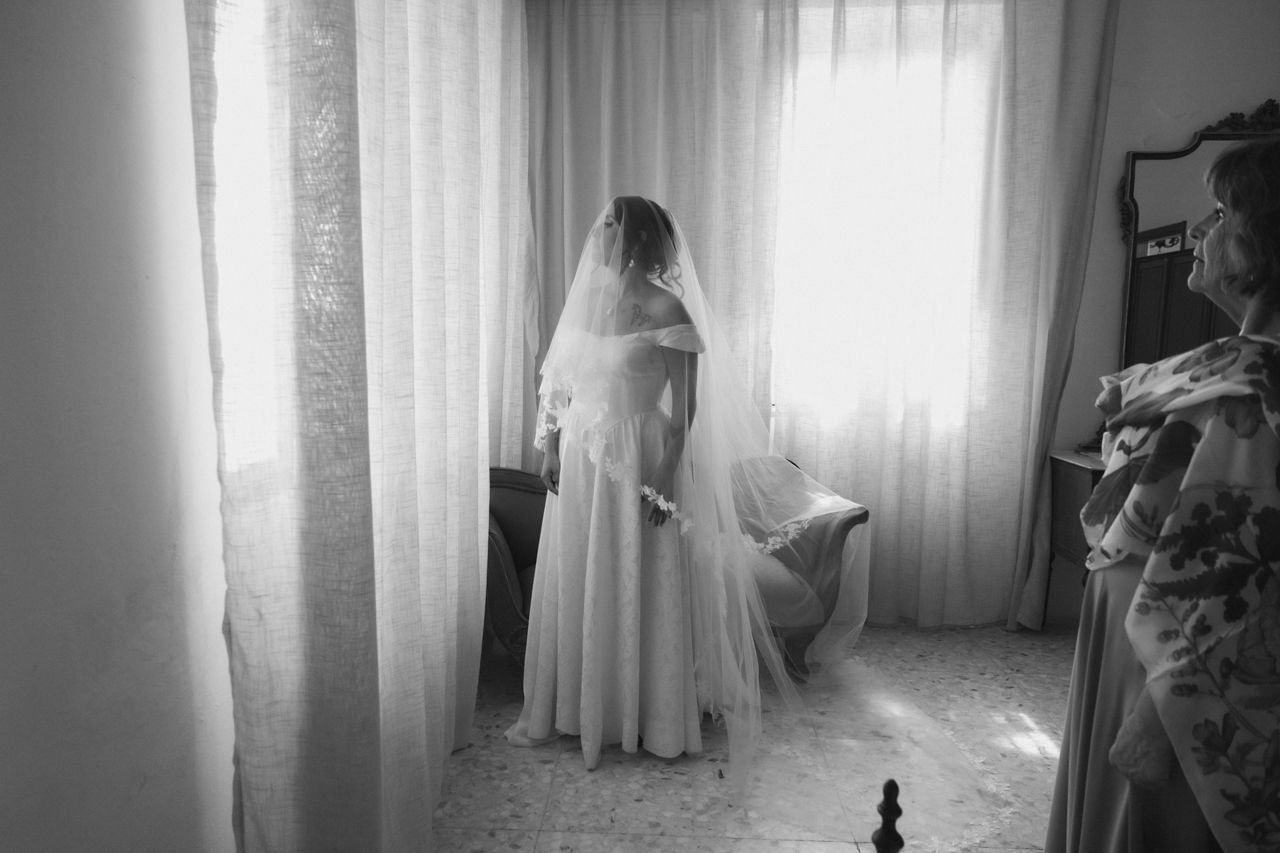 Black and white photo of bride in flowing dress preparing by window light.
