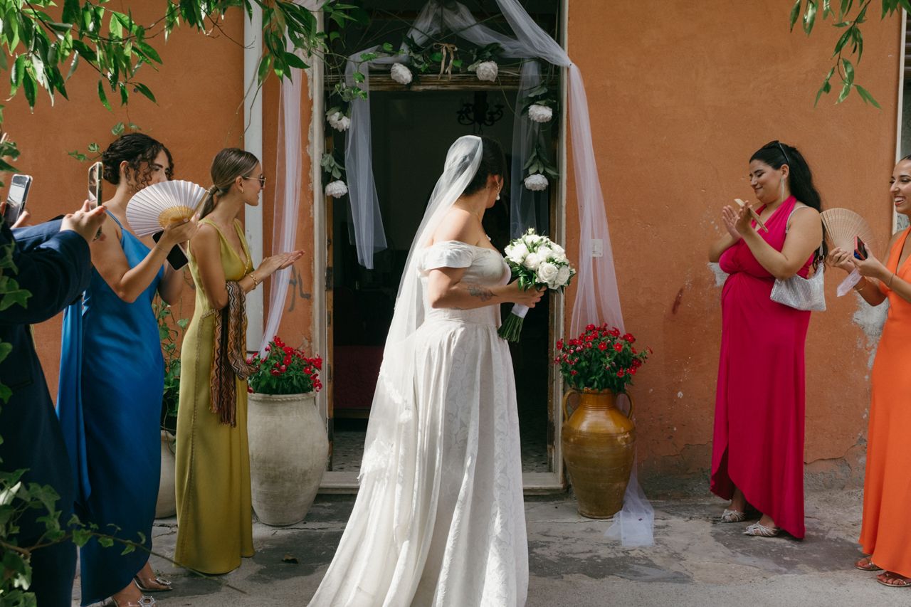 Wedding scene with colorfully dressed guests taking photos of bride outside venue entrance.