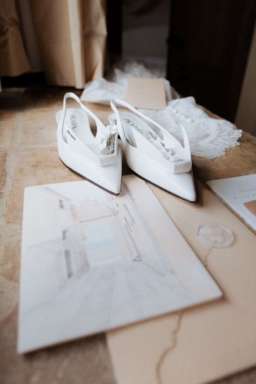 Elegant white pointed-toe bridal shoes beside wedding stationery on a marble surface.