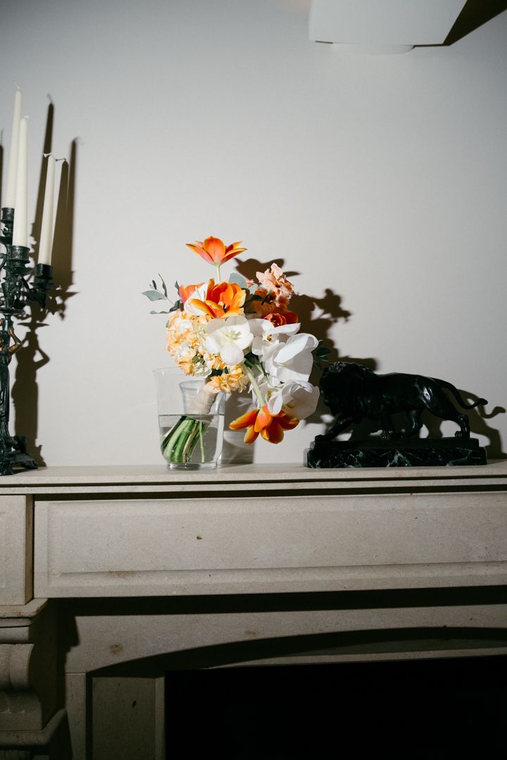 Orange and white floral arrangement in a glass vase with black candles on a sideboard.