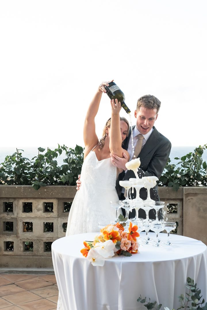 Wedding celebration scene with champagne tower and orange floral decorations on outdoor terrace.
