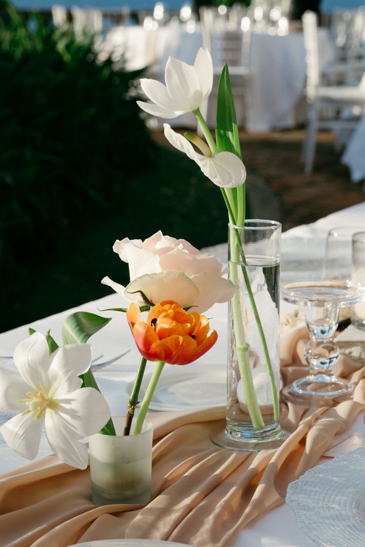 White and orange floral arrangements in glass vases on a champagne-colored tablecloth.