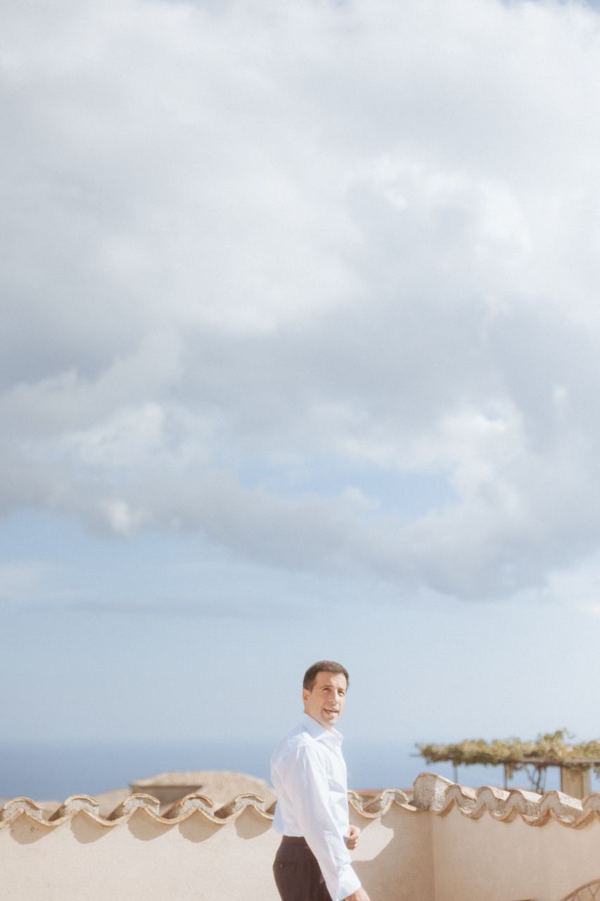 A person in white stands against a bright cloudy sky beside stone architectural elements.