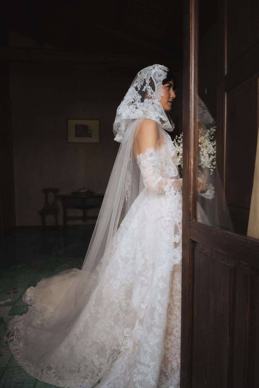 A side profile of an elegant bride in a lace wedding gown with flowing train and veil.