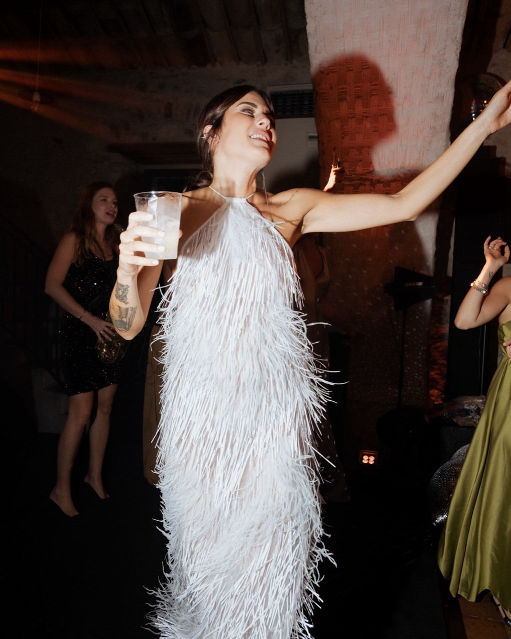 Woman in white fringe dress dancing at reception with drink in hand.
