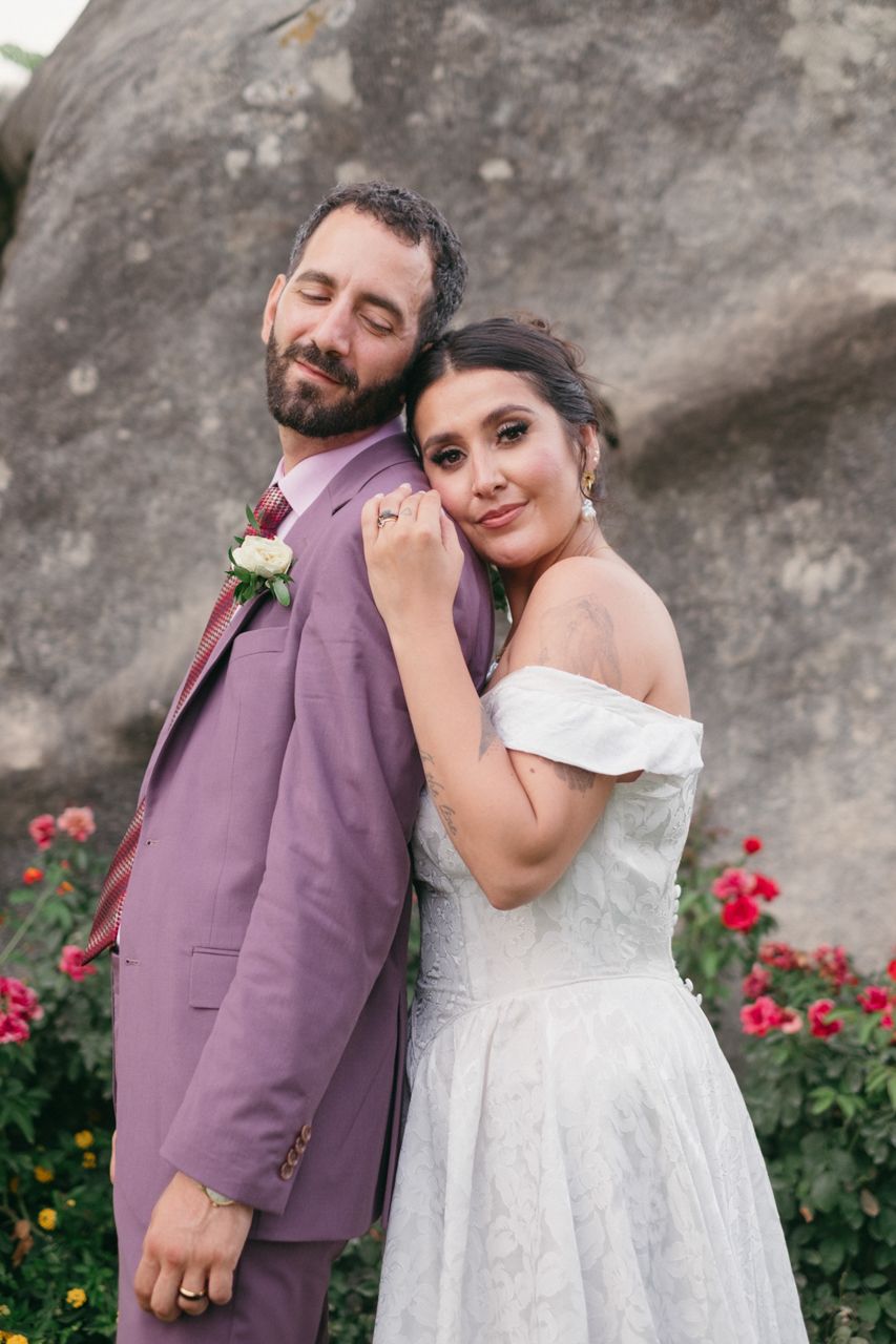 Romantic portrait of newlyweds in purple suit and white dress against floral garden backdrop.