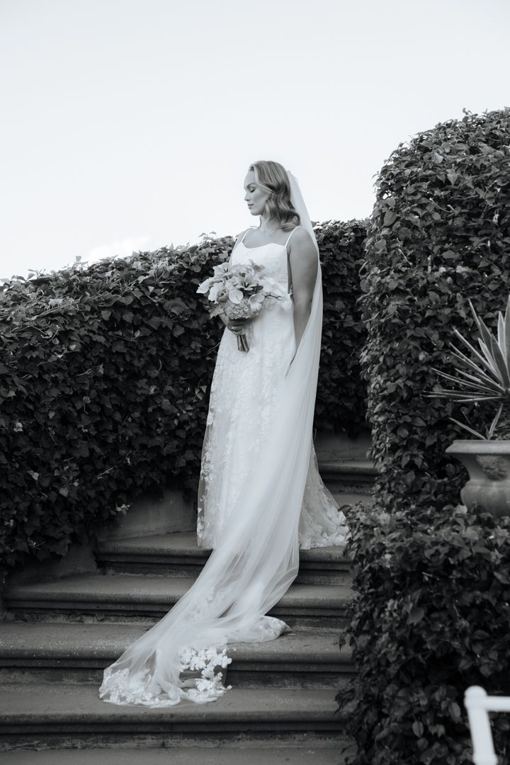 Bride in flowing white gown stands on garden steps holding bouquet against green hedge background.