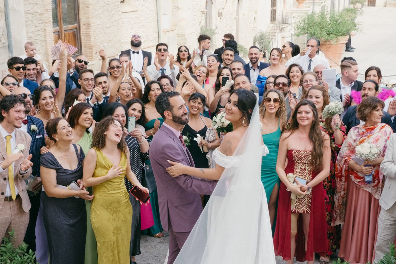 Large wedding gathering with guests celebrating newlyweds in front of stone building.