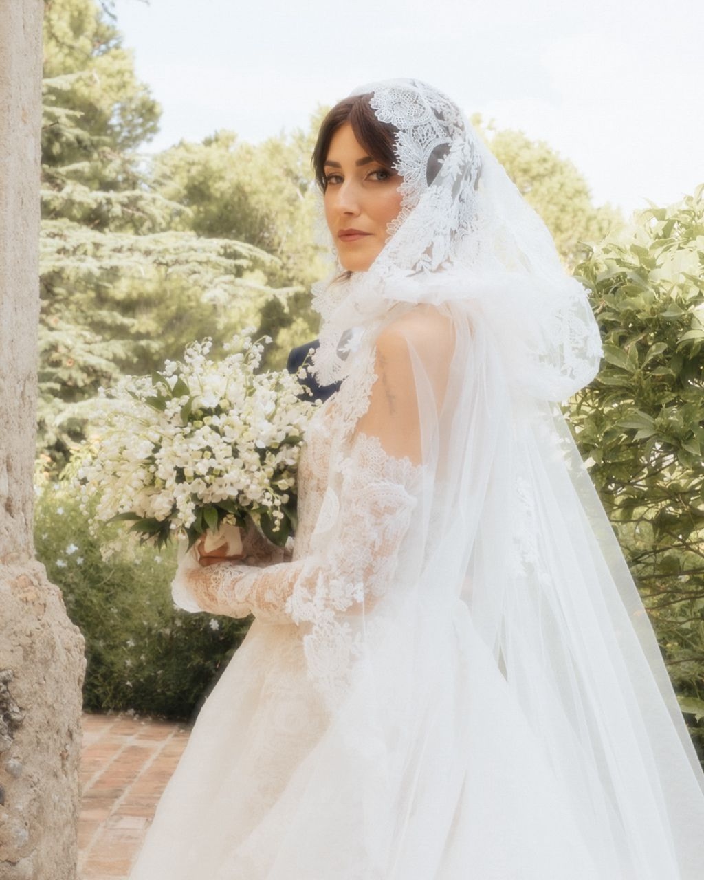 Bride in lace wedding gown and veil holds white floral bouquet.