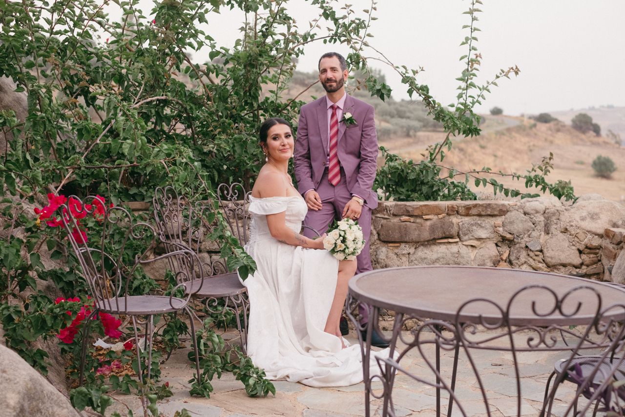 A wedding couple poses together on an elegant stone patio with red flowers in the background.