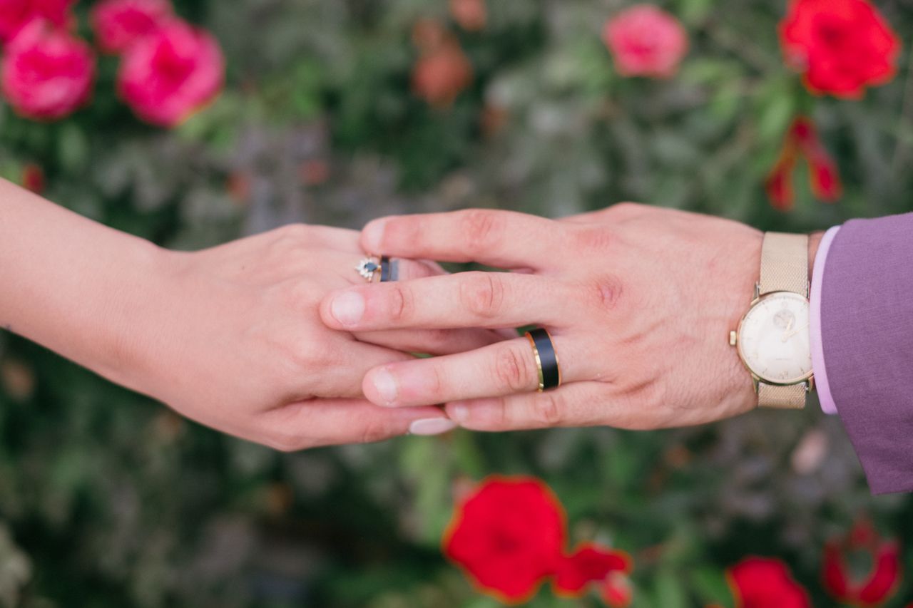 Close-up of hands with wedding rings against backdrop of vibrant red roses.