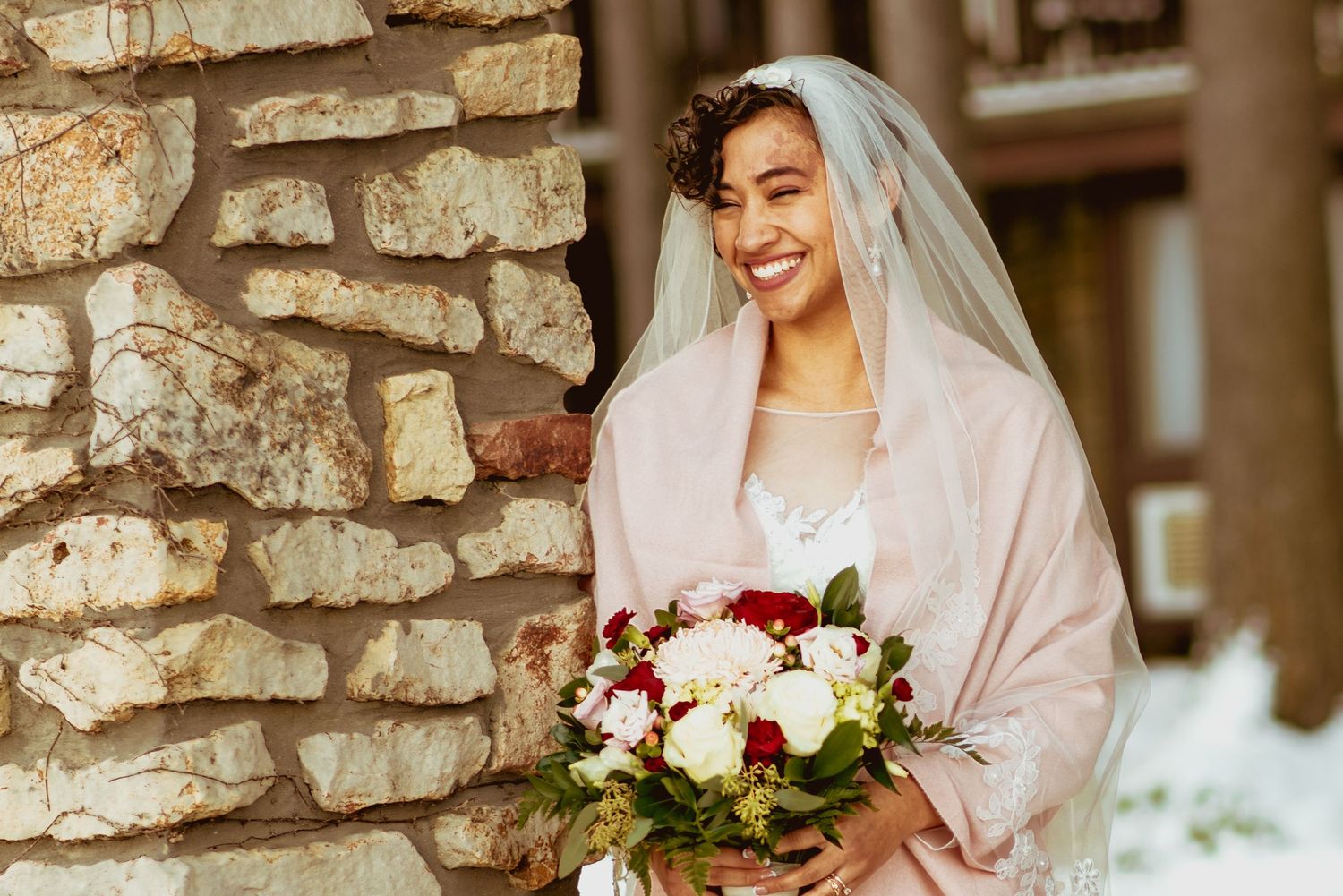 Bride in white dress poses with elegant bouquet featuring blush and cream colored flowers against rustic stone wall.