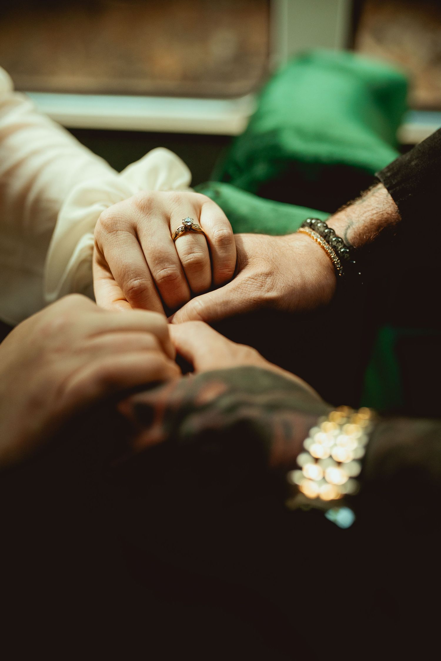 A close-up shot of hands clasped together showing a gold ring and bracelet in dim lighting.