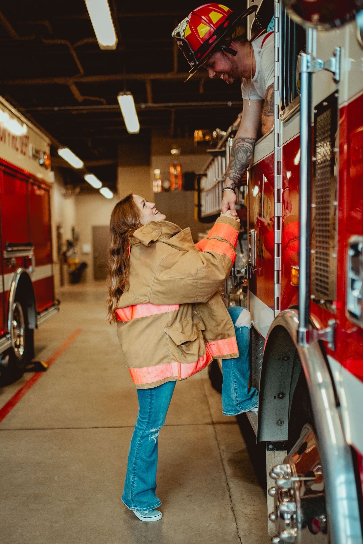A couple in firefighting gear share a romantic moment on a fire truck in a station bay.