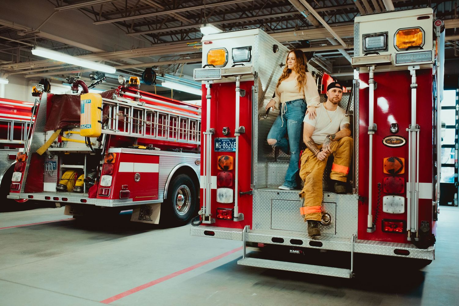 Two people in firefighting gear stand on a fire truck in the station garage.