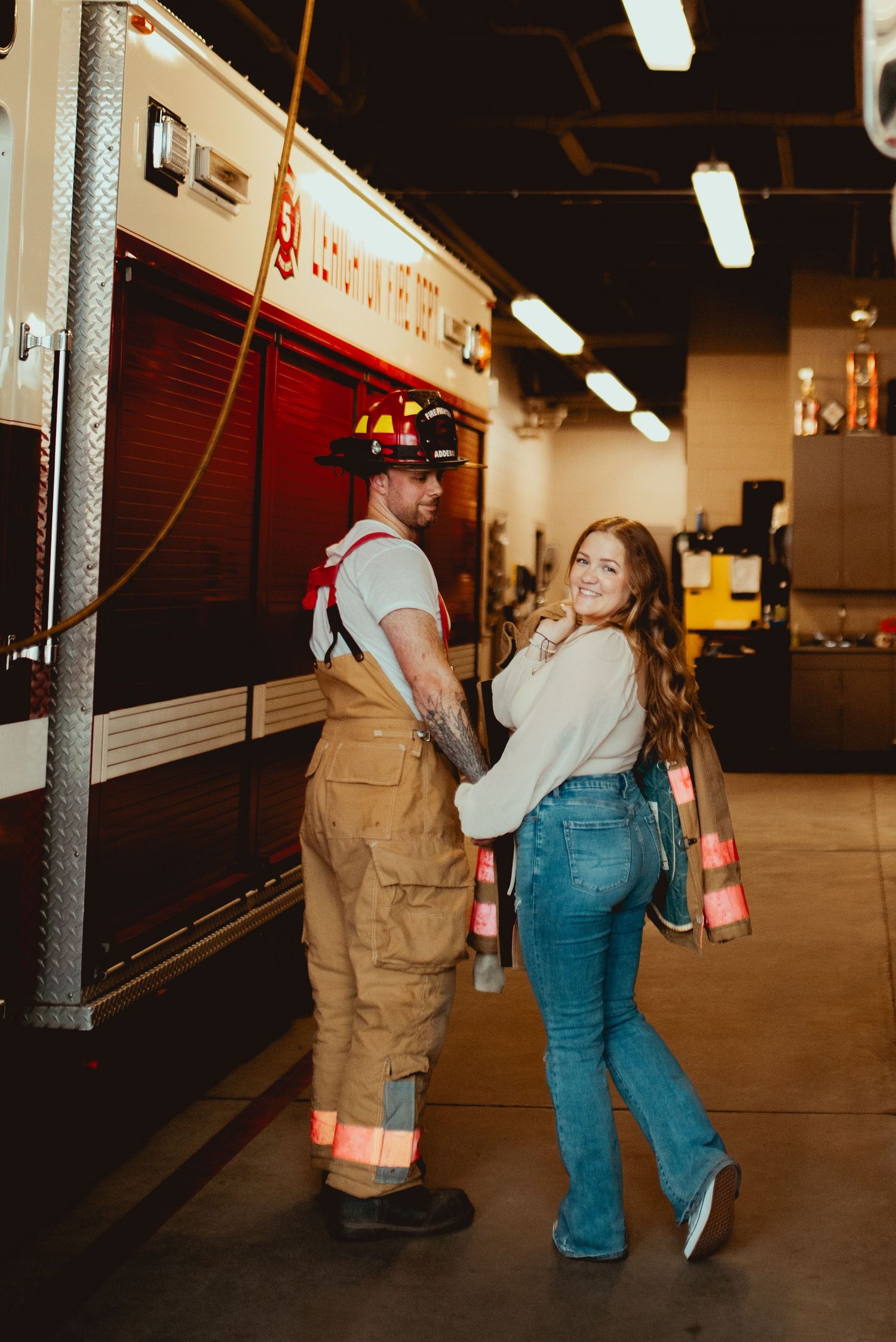A couple in firefighting uniform poses playfully next to a fire truck in the station.