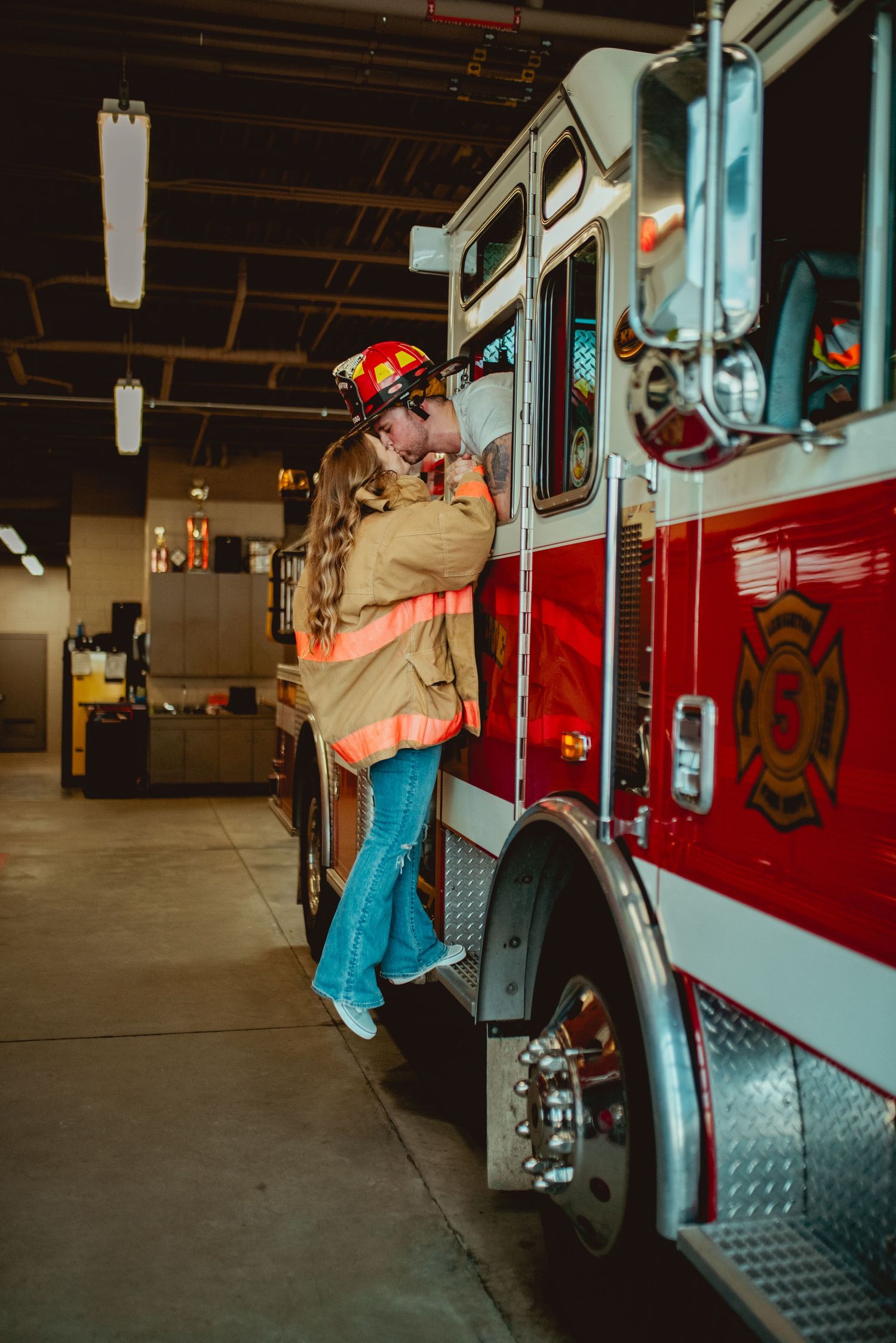 A romantic embrace between two people by a fire engine in the station garage.
