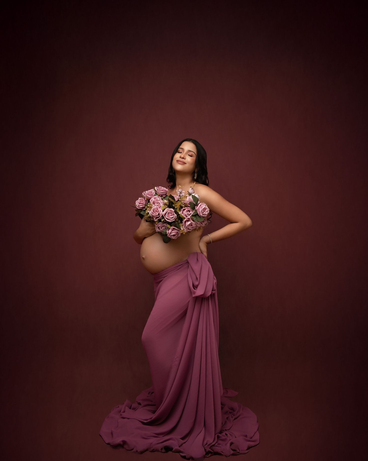 An elegant maternity photo features a woman in a flowing mauve dress holding pink and white roses against a burgundy backdrop.