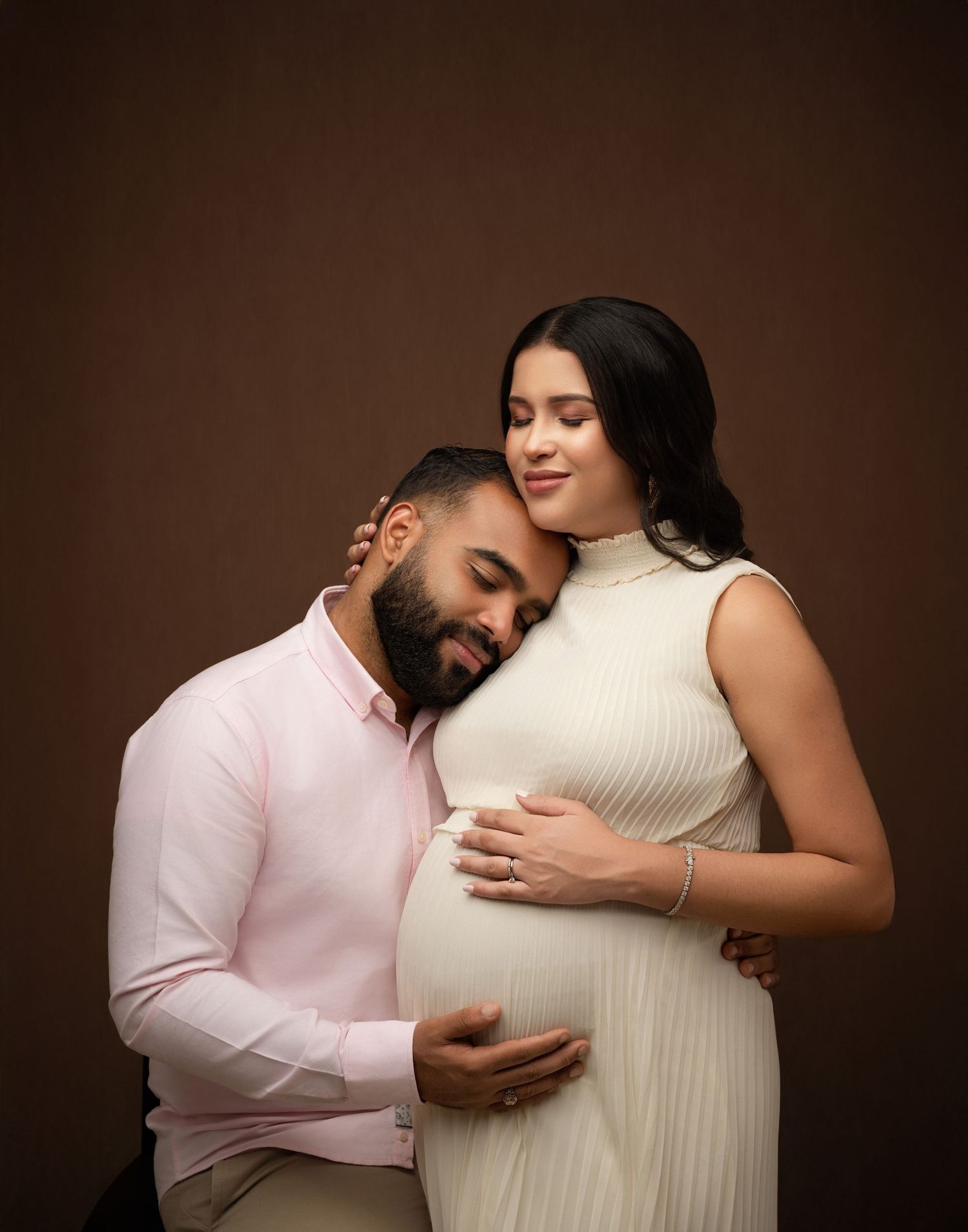 Intimate maternity portrait featuring a couple embracing against a brown backdrop with the partner resting head on pregnant belly.