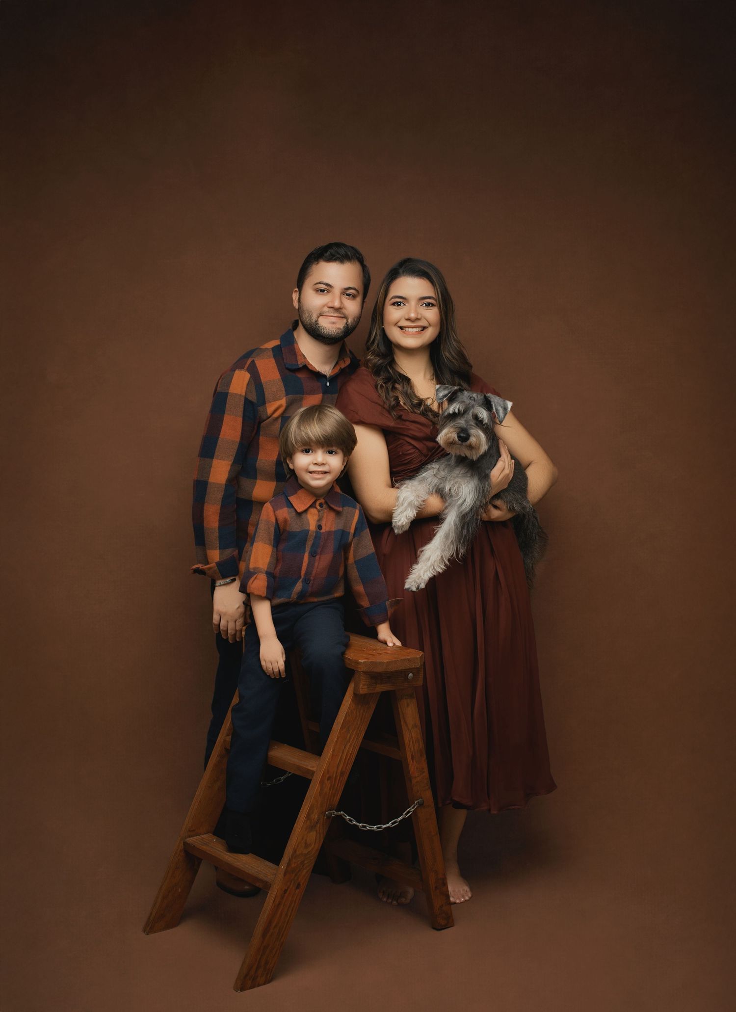 Family portrait in rustic style with plaid matching outfits, wooden stool, and small grey dog against brown studio backdrop.