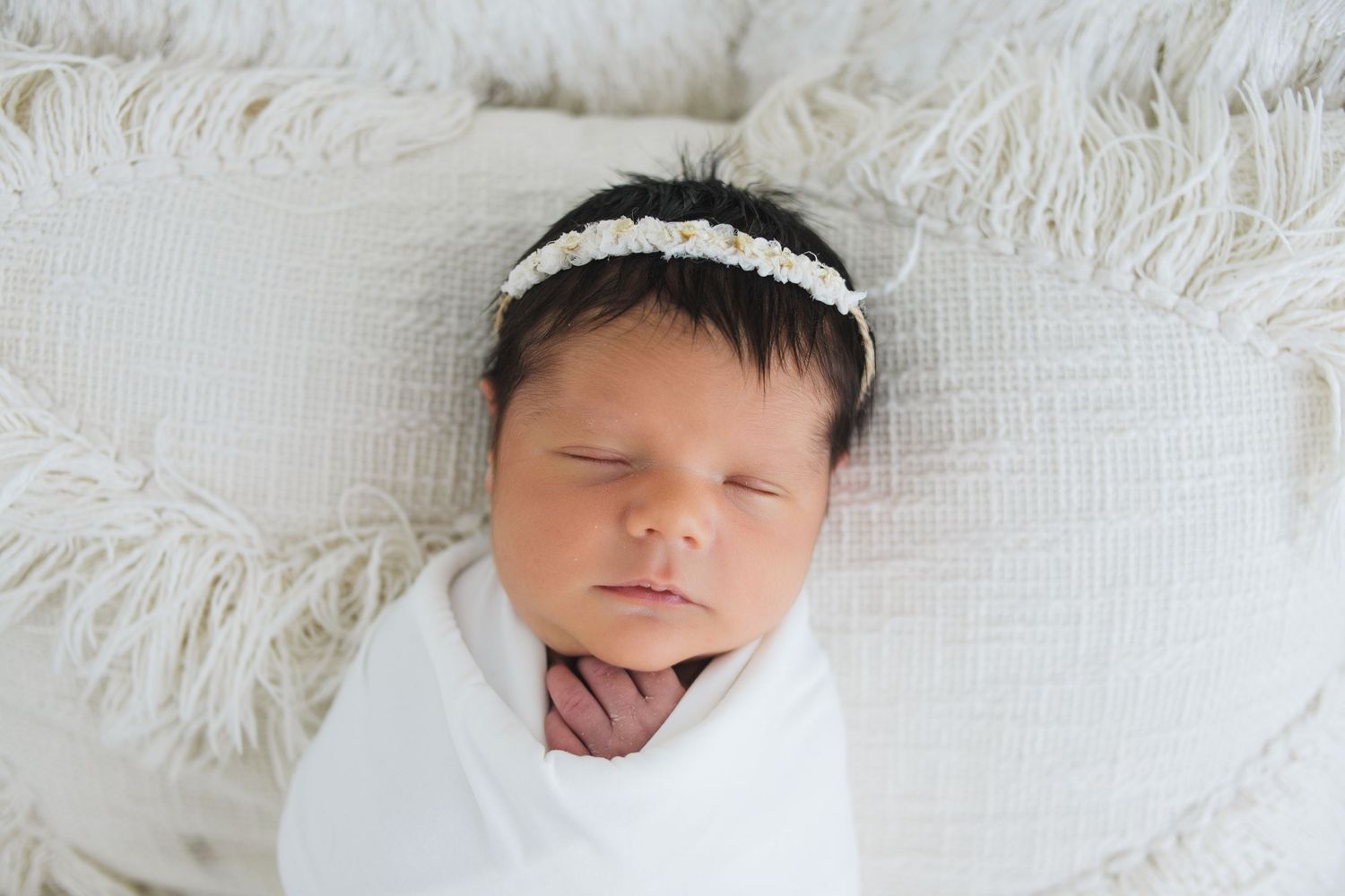 Peaceful sleeping newborn swaddled in white wrap with lace headband on soft white textured blanket.