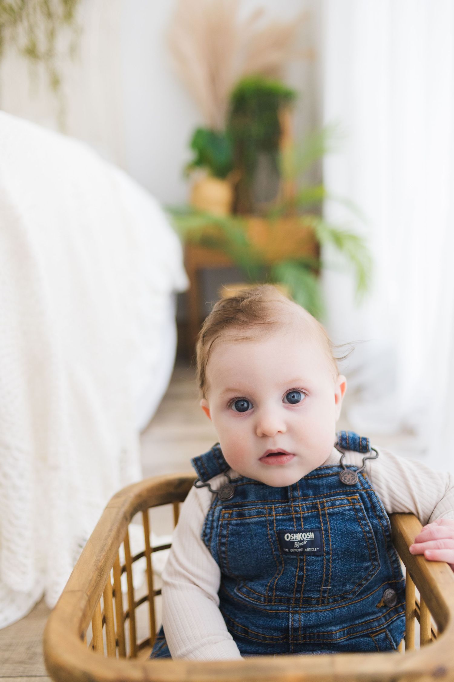 A baby wearing denim overalls sits in a woven rattan chair with plants in the background.