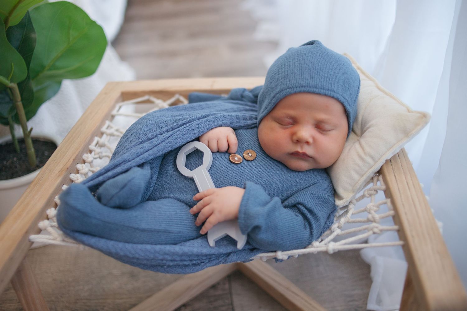 Sleeping newborn in blue outfit lying on wooden prop bed with white fabric and green plant.