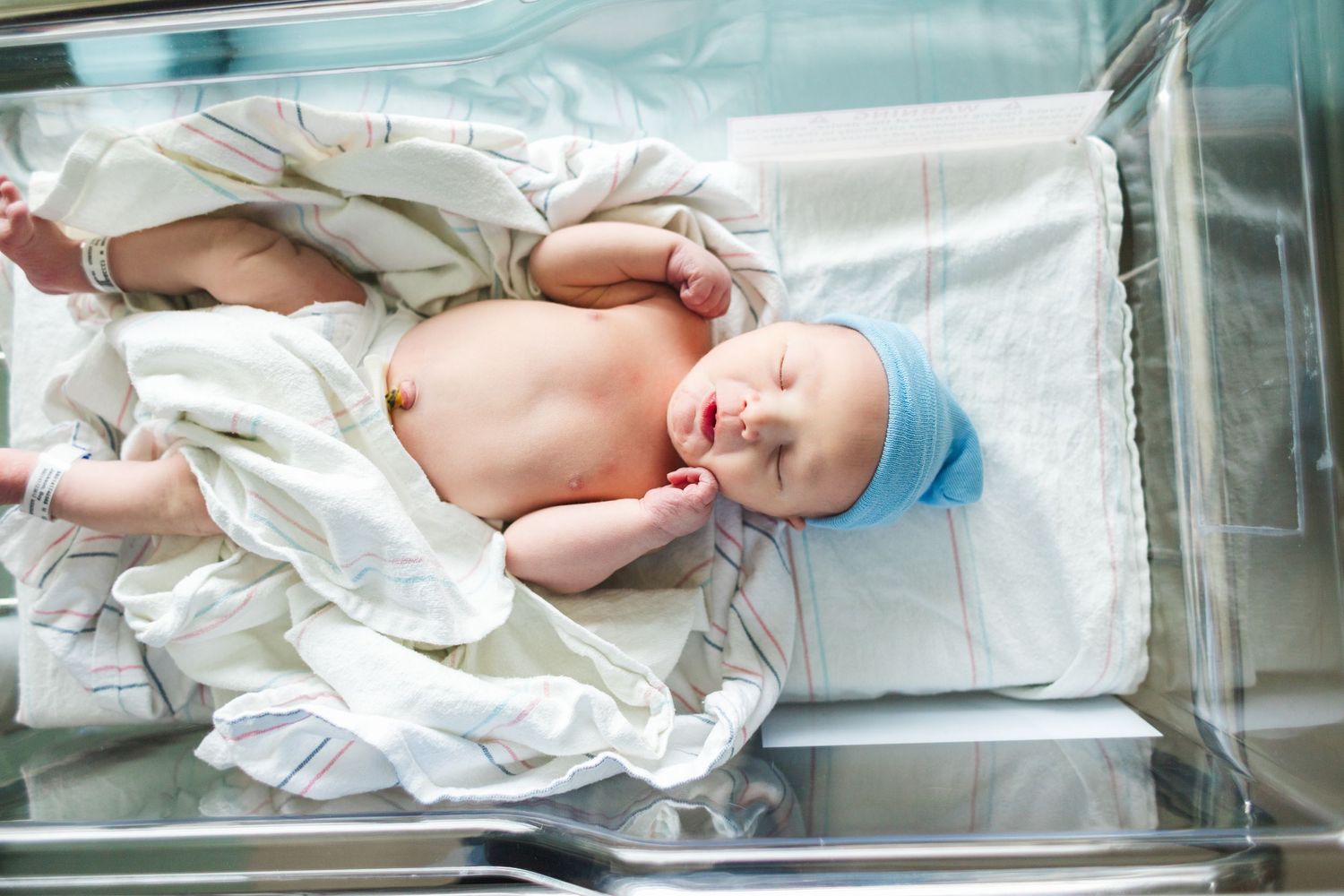 Newborn infant resting in clear hospital bassinet wearing white cloth and blue cap.