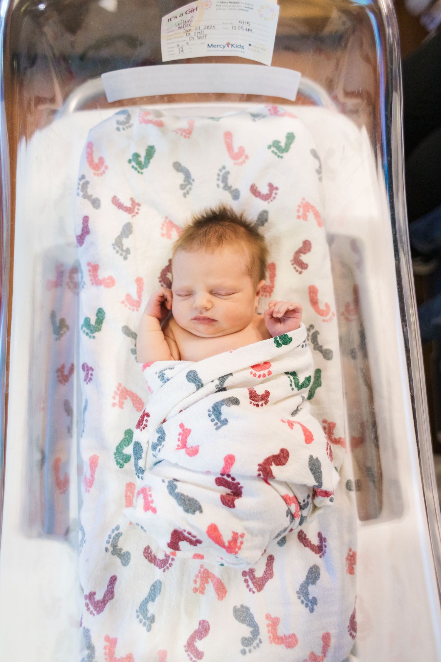 Sleeping newborn wrapped in footprint-patterned swaddle blanket in clear hospital bassinet.