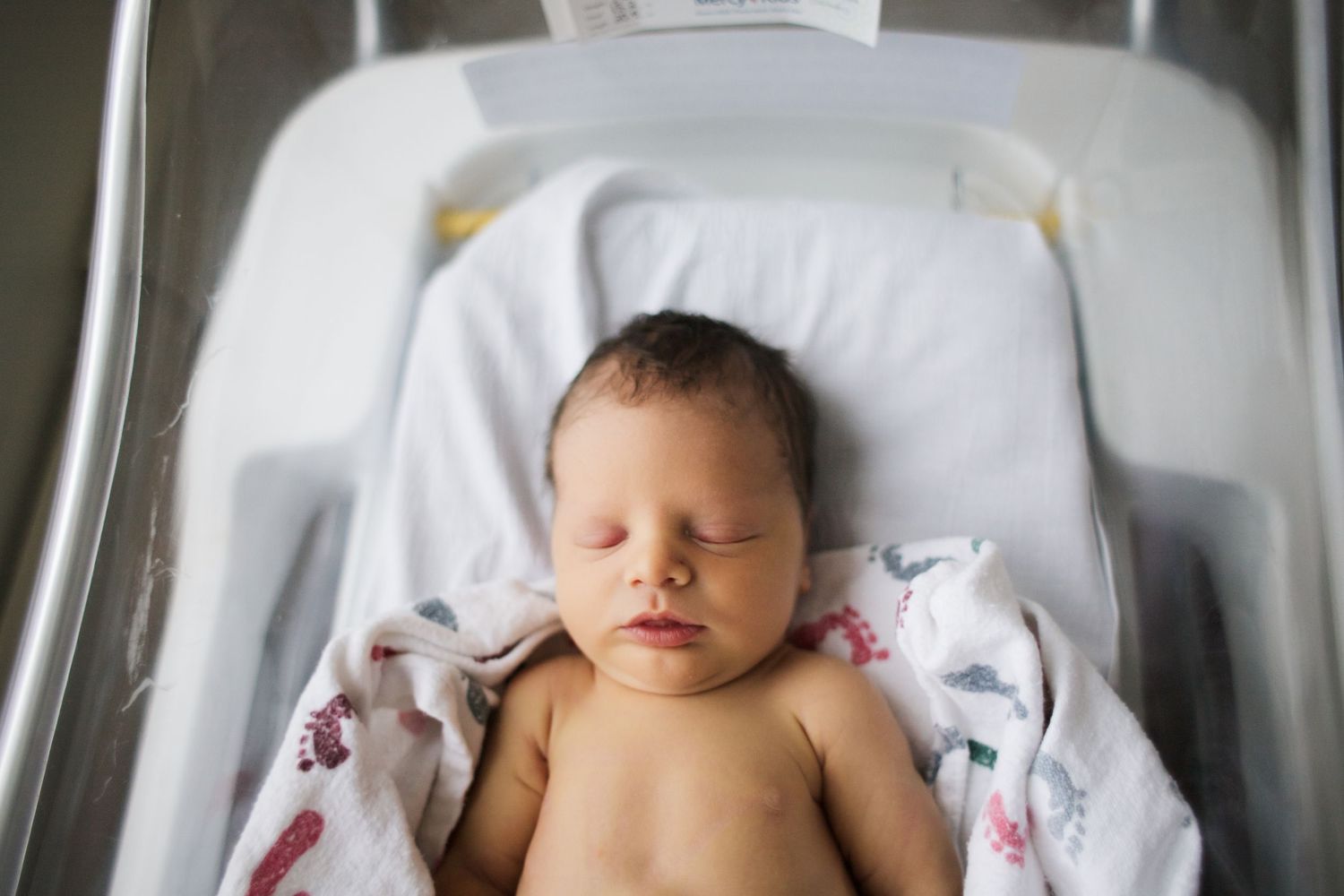 Newborn resting in hospital bassinet with white blanket and pillow.