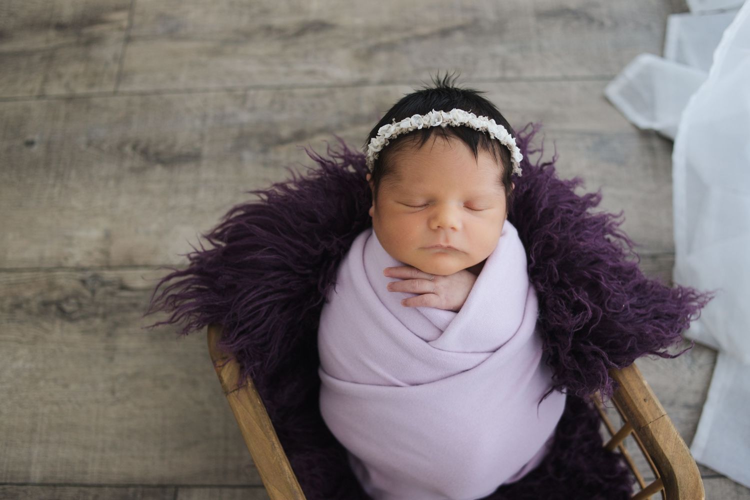 Sleeping newborn baby swaddled in soft pink wrap with purple feather decoration on wooden surface.