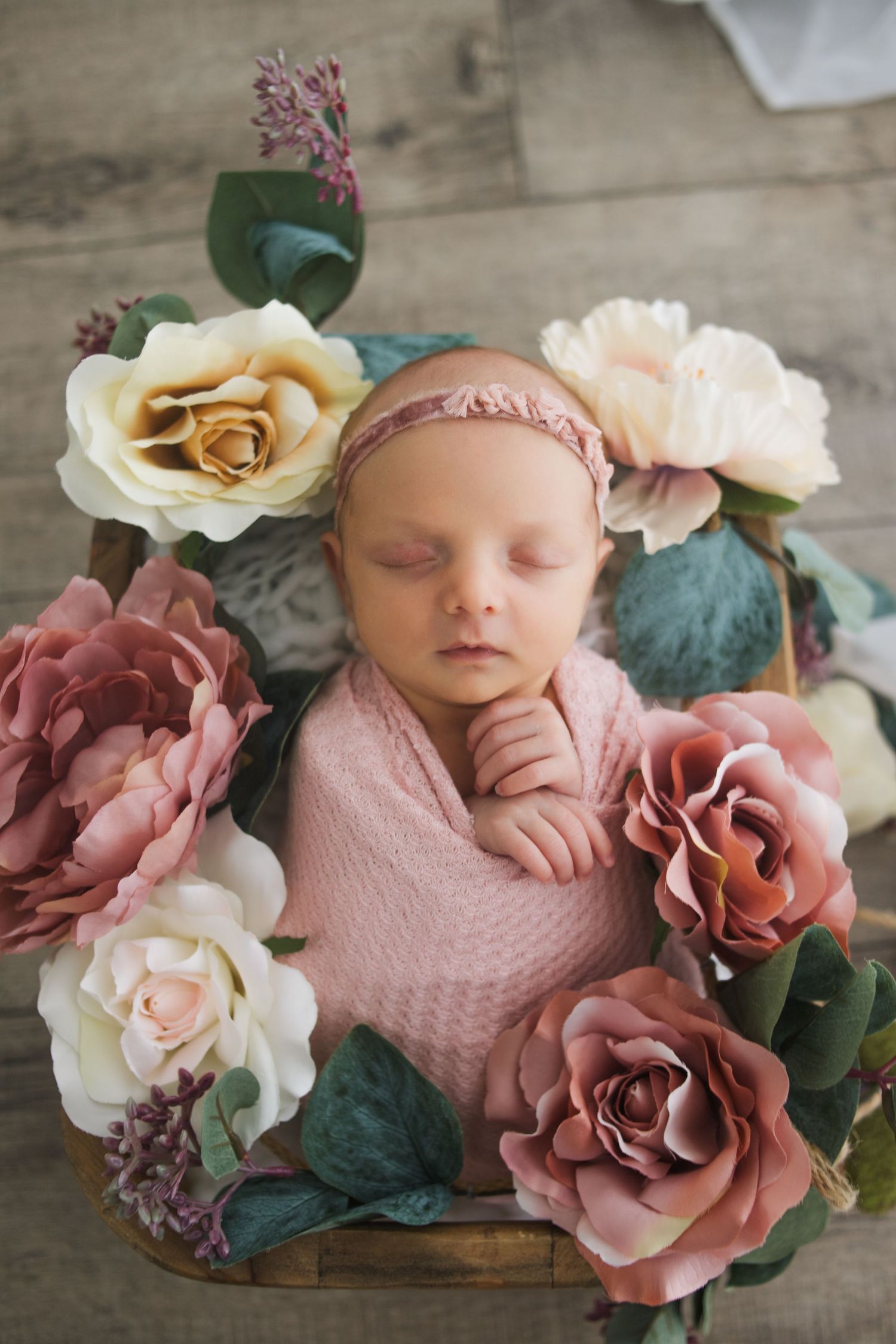 Newborn baby peacefully sleeping in pink swaddle surrounded by delicate dusty rose and cream artificial flowers on wooden surface.