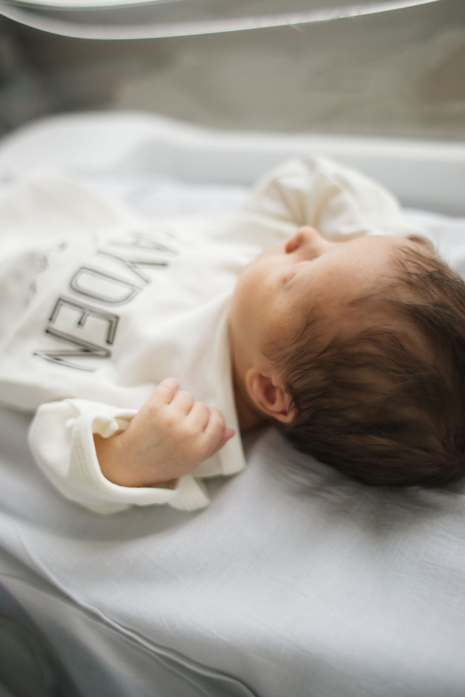 Close-up of sleeping baby wearing white clothing in hospital bed.