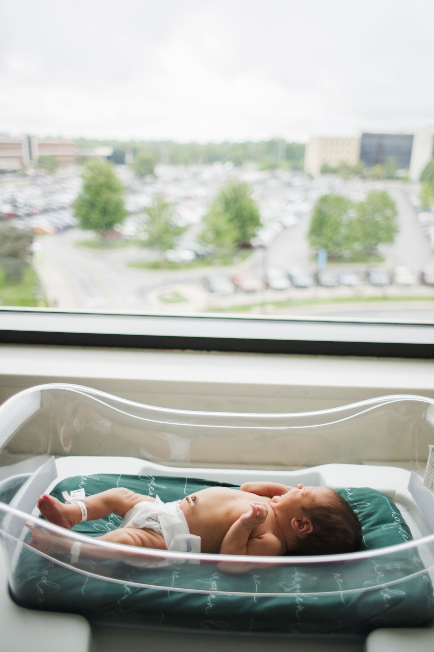 Newborn baby resting in clear hospital bassinet near window overlooking parking lot.