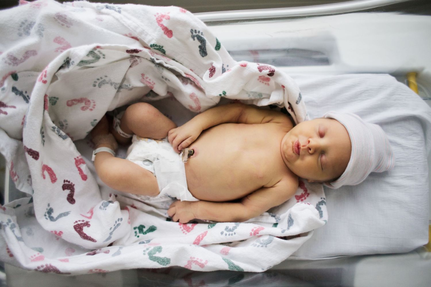 Newborn infant lying on white hospital bedding wrapped in patterned swaddle blanket.