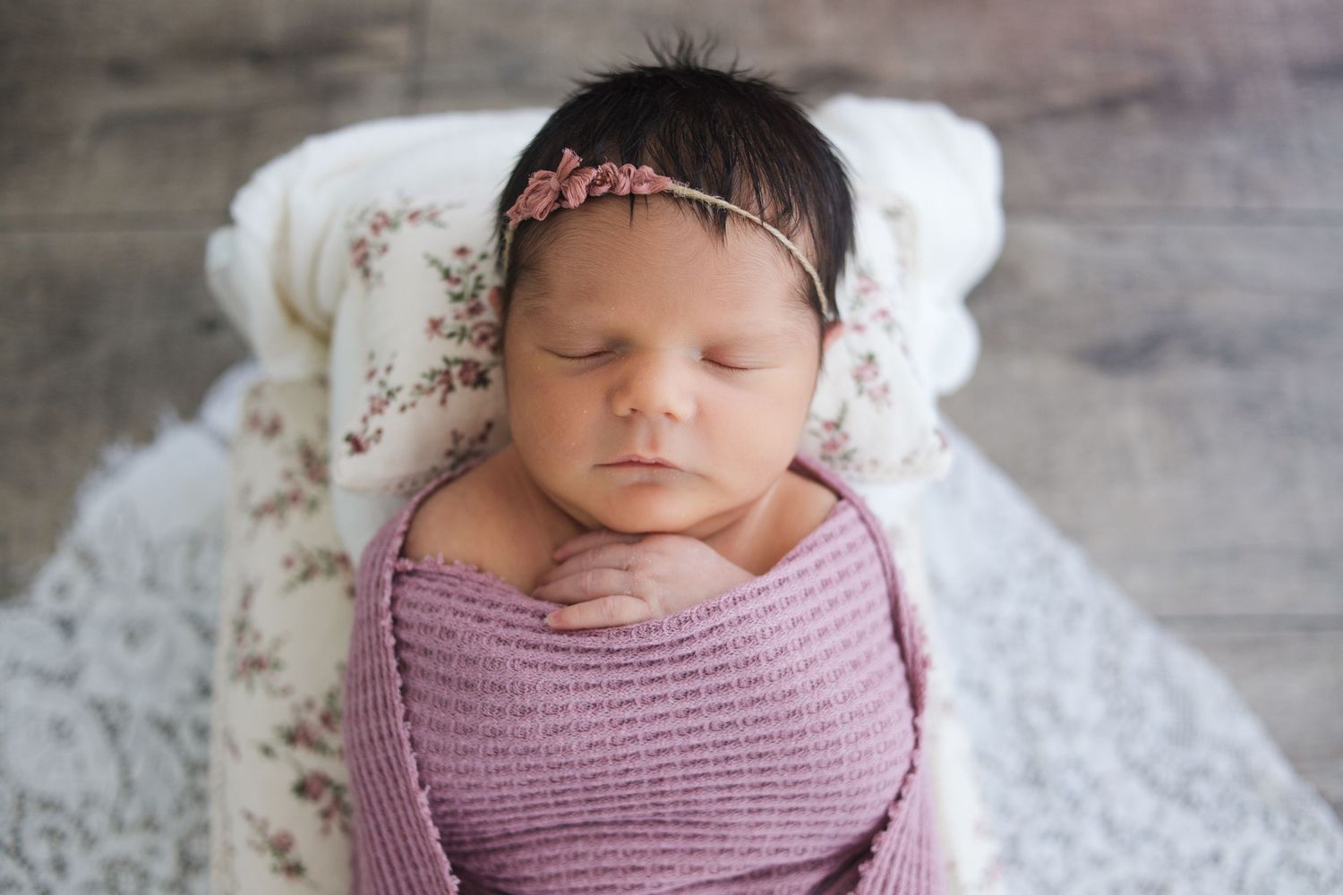 Sleeping newborn baby swaddled in mauve fabric against a soft lace backdrop with rustic wooden flooring.