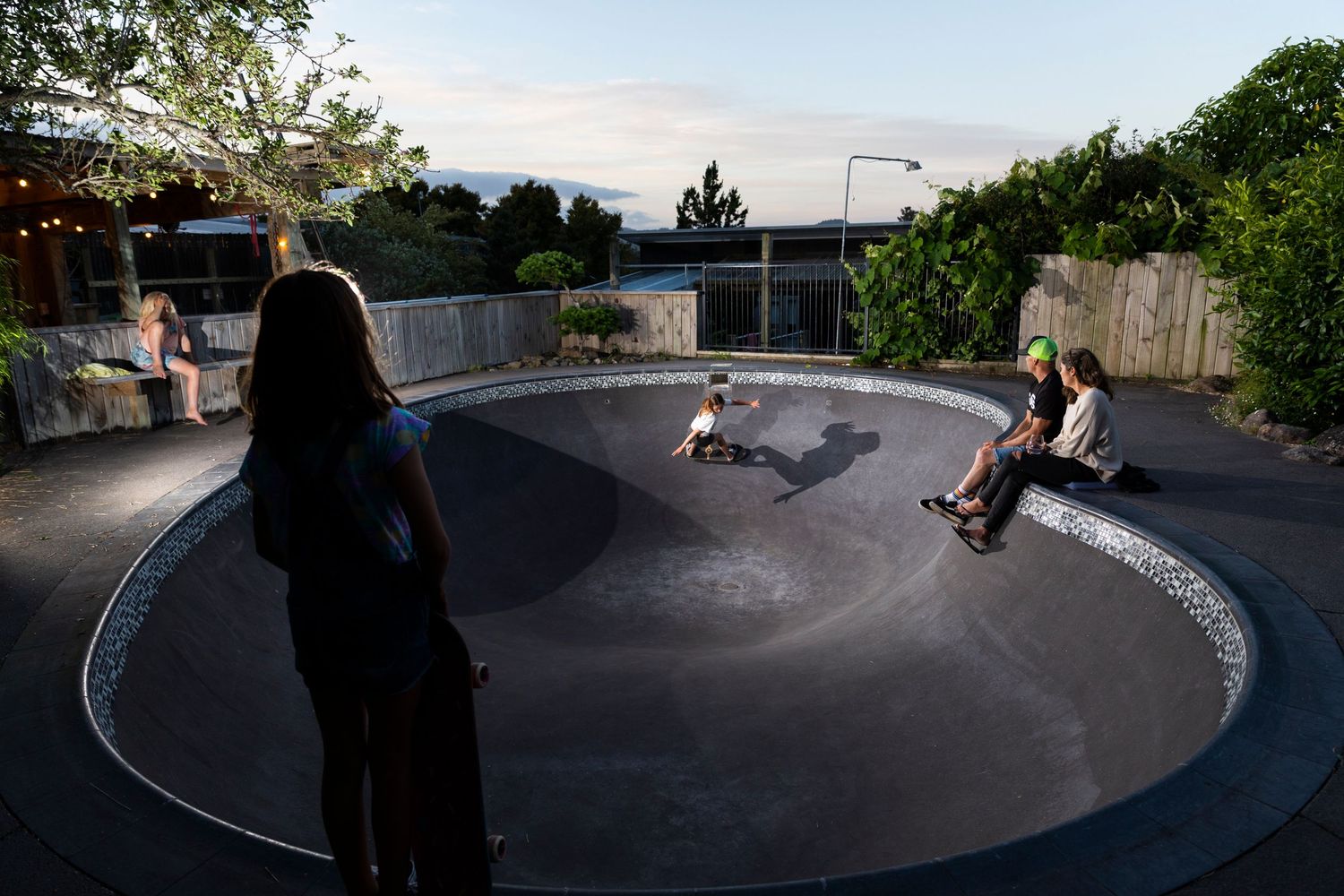 Skateboarders performing tricks in a large concrete bowl at dusk.