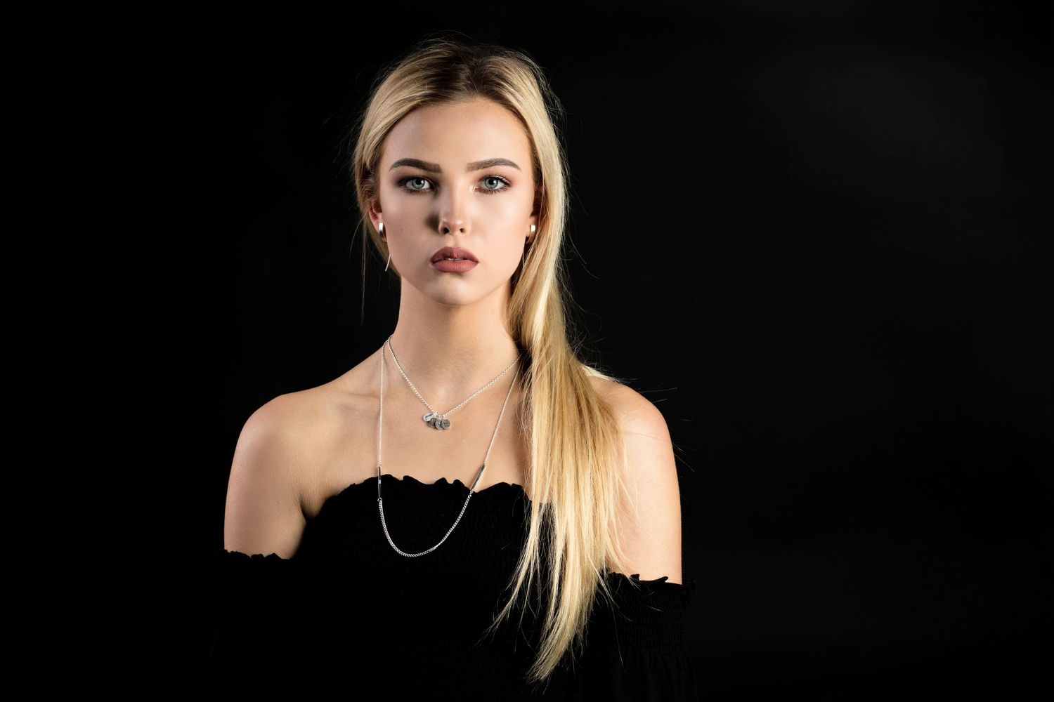 Dramatic portrait of person in black off-shoulder top with layered necklaces against dark backdrop.